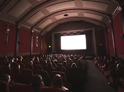 Darkened movie theater with lit-up screen. The backs of people's heads are visible above the set backs.