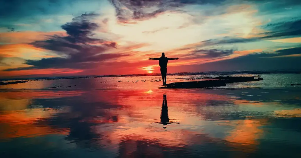 A silhouetted figure standing with outstretched arms standing on a beach at sunset