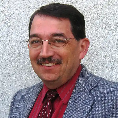 A middle-aged Caucasian man wearing a jacket, tie, and glasses