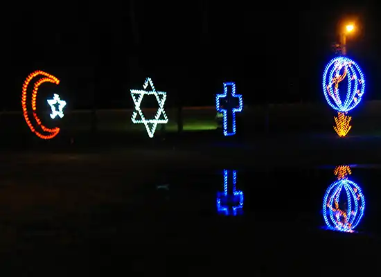 A Muslim crescent, Jewish star, and Christian cross outlines created with colored holiday lights. The shapes and colors reflect in the water below.