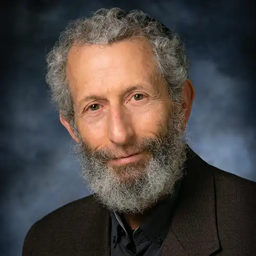 An older man with grey hair and beard wears a dark collared shirt and dark blazer