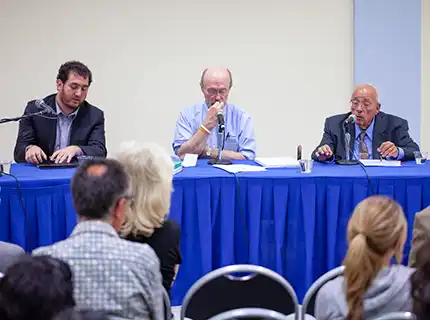 3 men seated at a table in front of an audience. Each man has a microphone in front of him and papers on the table.