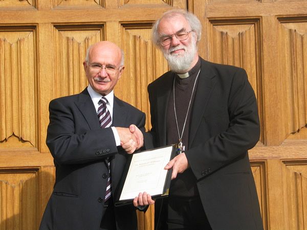 Two senior gentlemen shaking hands and holding a plaque while facing the camera. One man wears a suit and tie. The other wears clerical robes.