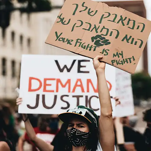 Female protest marcher holding up a sign written in Hebrew. Behind her is someone holding up a sign that says We Demand Justice.