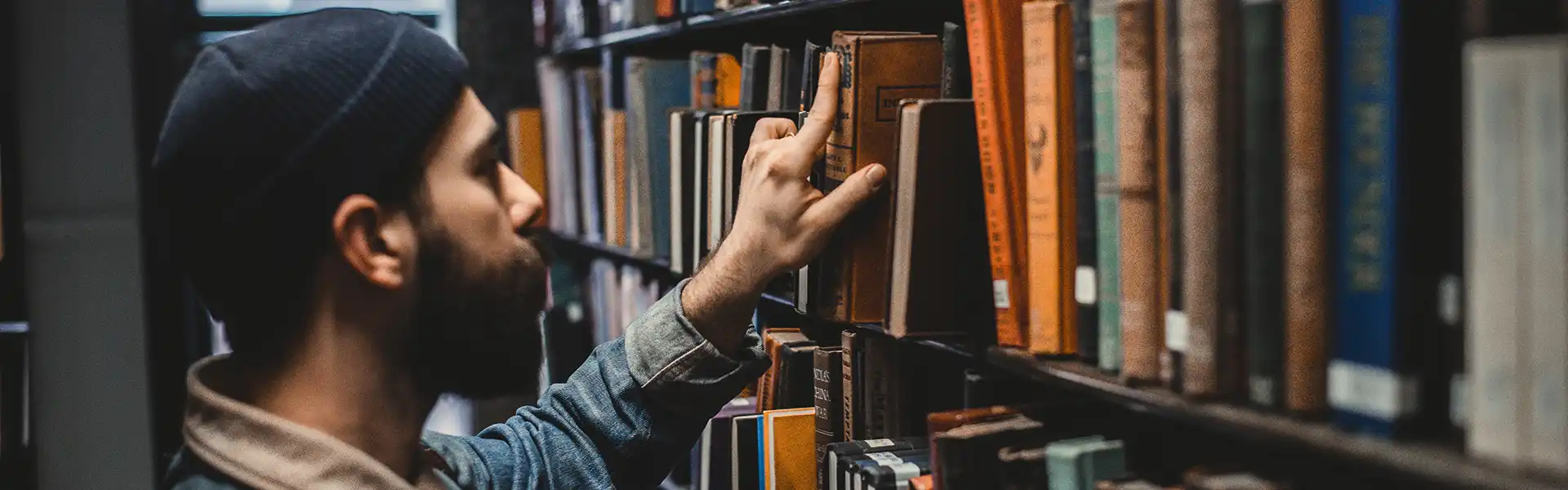 Man with a beard wearing a knit cap and denim jacket pulls a book off the shelf in a library