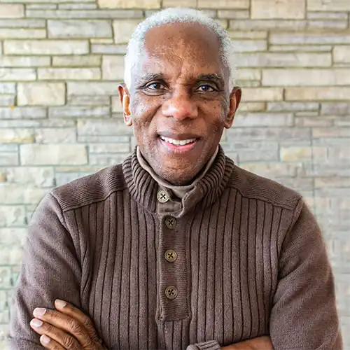 An older black man with white hard stands in front of a stone wall wearing a brown sweater. His arms are folded in front of his chest.