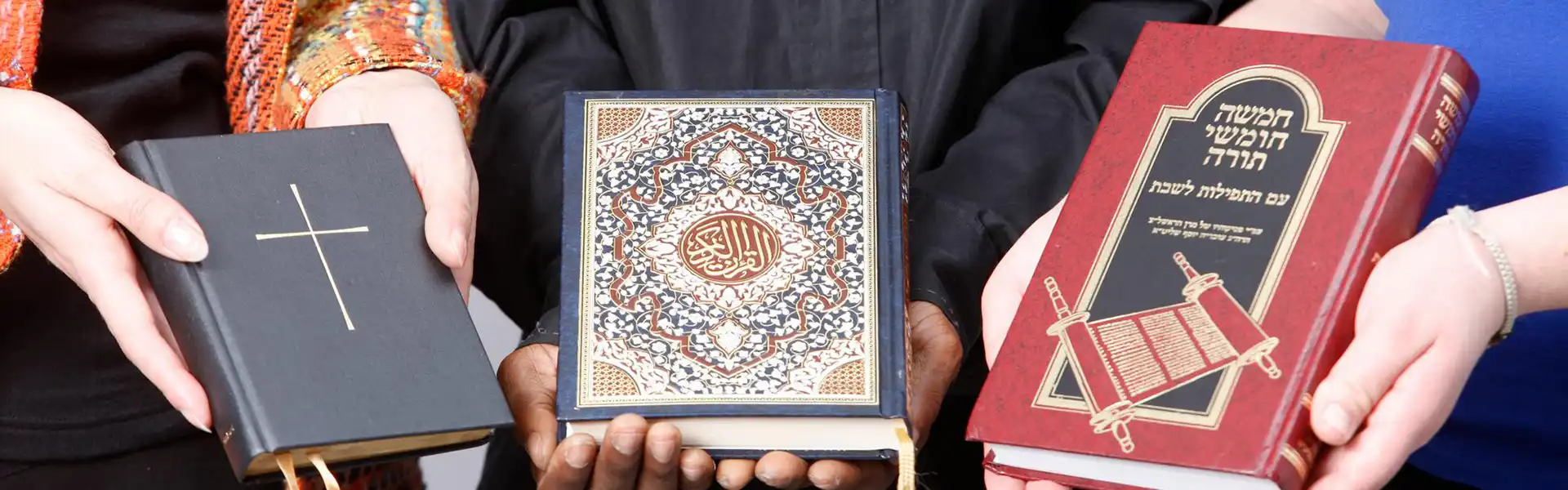 A close-up on the hands of 3 people holding a Christian bible, the Quran, and the Torah
