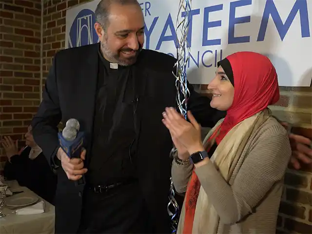 Father Khader El-Yateem holds a microphone and looks over at an Arab-American woman standing next to him. She wears a traditional headscarf. They stand in front of a campaign sign.