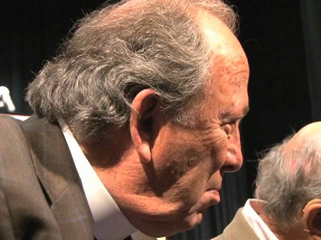 An older man leans into a conversation on stage.